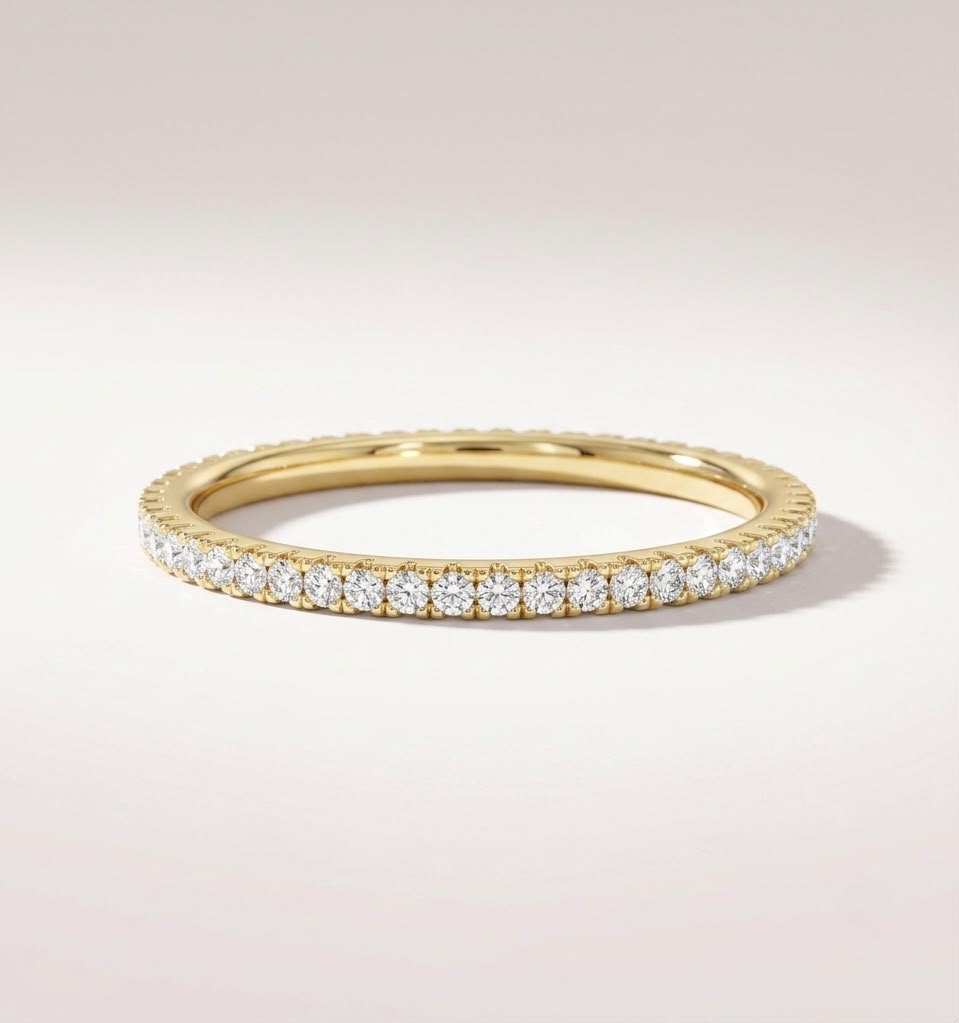 A yellow gold eternity ring set with round brilliant-cut diamonds, shown on a clean, light beige background.