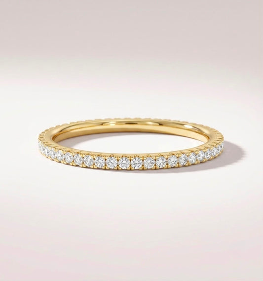 A yellow gold eternity ring set with round brilliant-cut diamonds, shown on a clean, light beige background.